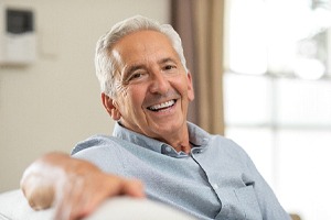 An older gentleman sitting on a couch and smiling to show off his dental implants after undergoing a bone graft
